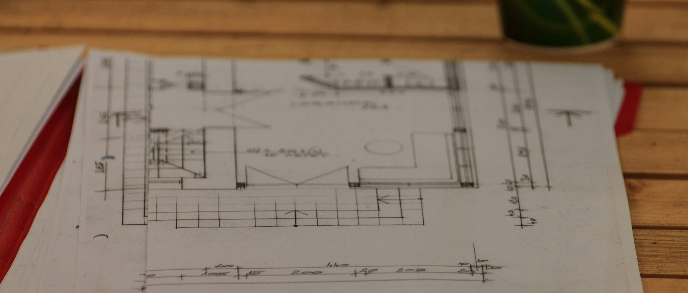 Handgezeichneter Grundriss mit Maßketten auf einem Stapel Planunterlagen in einer roten Mappe, auf einem Holztisch liegend